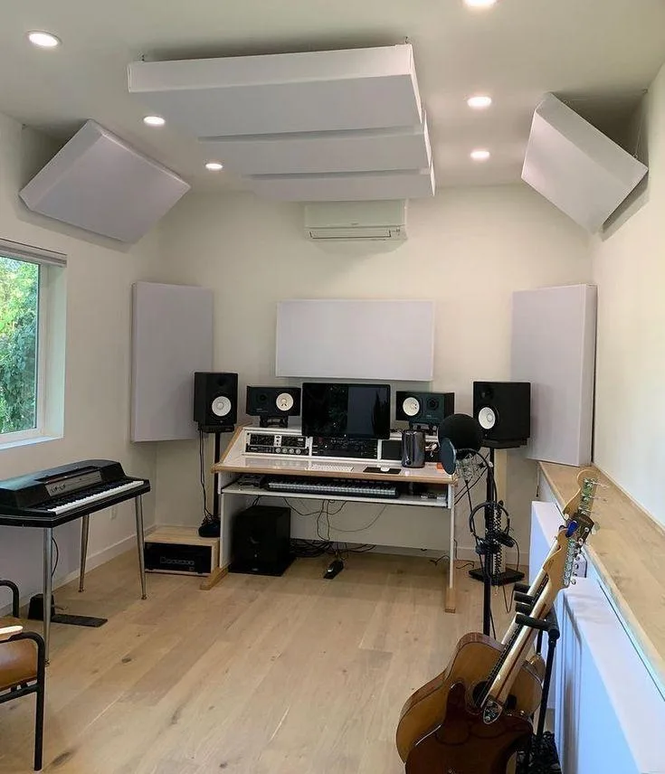 Estudio doméstico claro con varios paneles suspendidos del techo inclinado y un panel rectangular en la pared, tras un escritorio con cuatro monitores; a un lado un piano digital y al otro una guitarra.